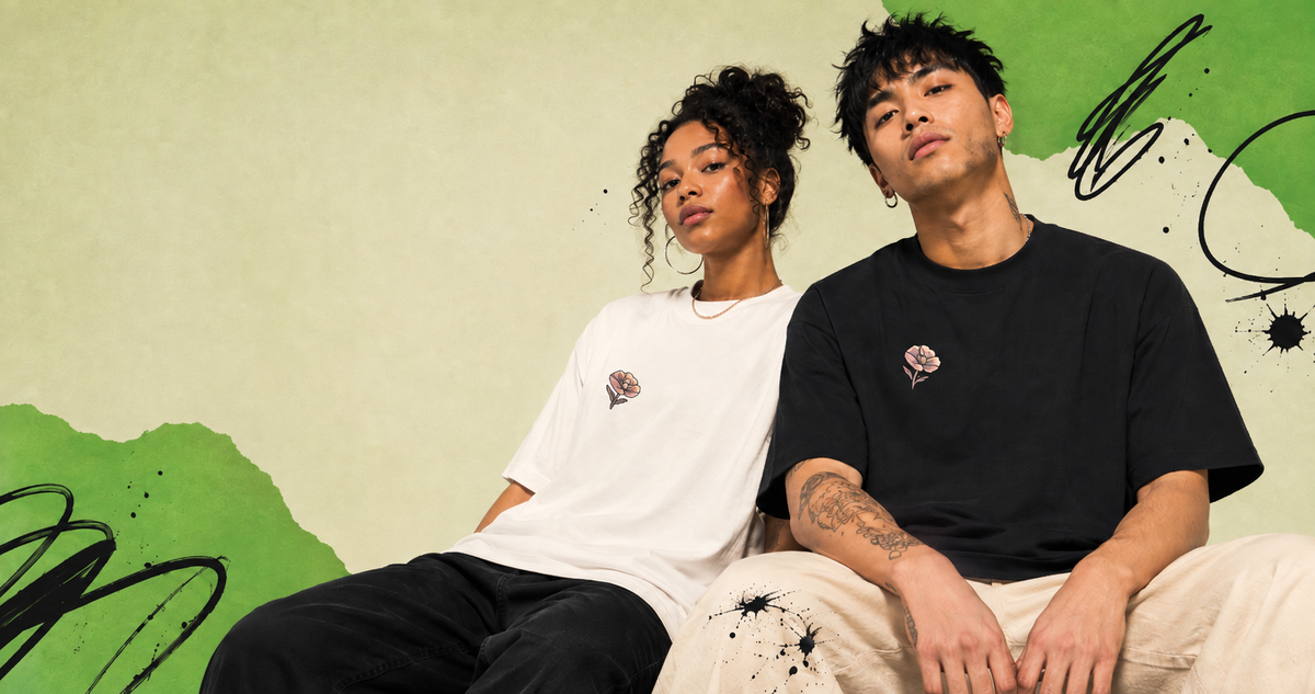 Two models sitting side-by-side wearing matching graphic t-shirts. The model on the left wears a white t-shirt with a small pink graphic on the left chest, paired with black pants.