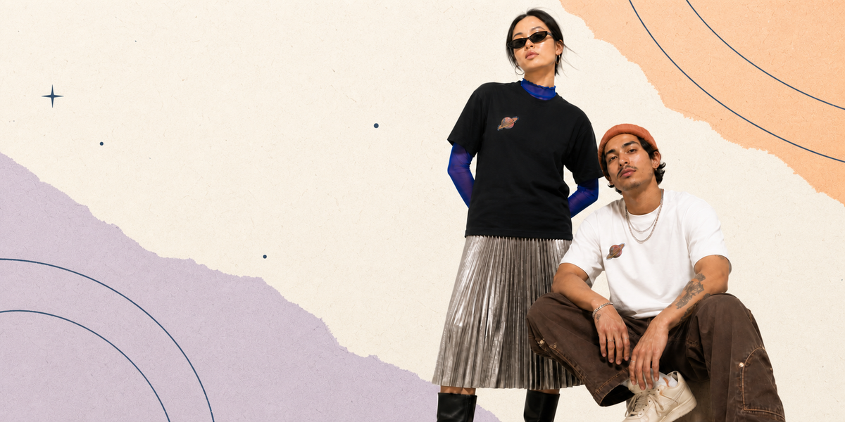 Two models posing in front of a stylized abstract background. The standing model wears a black crewneck t-shirt with a small chest graphic over a blue long-sleeve top, paired with a pleated metallic silver skirt.